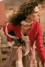 A woman in a vintage-style outfit wearing a red knit cardigan, white lace ruffles, a brown gingham skirt, and lace arm warmers, captured in a warm, rustic setting.