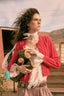 A woman outdoors wearing a bright red cable-knit cardigan over a white lace blouse, holding a small stuffed animal. The background shows rustic buildings and hills under a cloudy sky.