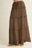 A rust-colored, tiered maxi skirt with a drawstring waist, shown on a model wearing light-colored sandals against a plain beige background.