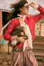A woman outdoors wearing a bright red knit cardigan over a white lace blouse, with a brown plaid skirt and holding a small stuffed animal.