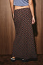 A person wearing a long dark plaid skirt with brown and red lines, paired with a blue crop top and black shoes, standing against a wood-paneled wall.