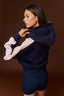 A woman wearing a navy blue color-blocked hoodie with light pink sleeves and matching navy shorts, posing against a solid dark brown studio background.