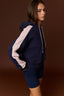 A woman wearing a navy blue color-blocked hoodie with light pink sleeves and matching navy shorts, posed against a solid dark brown studio background.