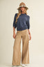 A woman modeling a navy blue knit sweater with three-quarter sleeves, paired with beige wide-leg trousers, a woven straw hat, and white sneakers, set against a plain beige studio background.