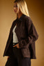 A woman wearing an oversized dark brown corduroy button-up shirt jacket over a white tank top and black jeans, posed against a warm brown studio background.