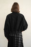 A person shown from the back wearing a black cropped bomber-style jacket with voluminous sleeves and a black-and-white plaid skirt, against a plain off-white wall.