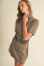 A woman wearing a taupe knit short-sleeve turtleneck dress with a matching wide belt featuring a gold buckle, posed against a solid light beige background.