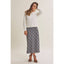 A woman modeling a white knit crewneck sweater paired with a navy and gray plaid midi skirt and black strappy high-heeled sandals, set against a plain light beige studio background.