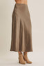 A taupe brown satin midi skirt worn by a model, paired with beige ankle boots, shown from the waist down against a light beige studio background.