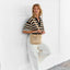 Woman wearing a striped shirt and white pants, holding a beige crossbody bag against a white wall.