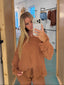A woman takes a mirror selfie in a fitting room wearing a matching rust-brown button-up shirt and pants set. The fitting room has light wood doors, a mirror, and clothing racks in the background.