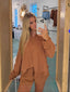 A woman takes a mirror selfie in a boutique, wearing a matching rust-brown lounge set consisting of a long-sleeve button-up shirt and shorts. The setting features light wood framing around the mirror, and clothing racks are visible in the background.