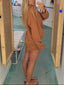 A woman takes a mirror selfie in a fitting room, wearing a loose, long-sleeve brown oversized shirt and shorts. The fitting room has light wood-framed mirrors, a teal floor, and clothing racks in the background.