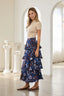 A woman stands in a bright white room with arched doorways and columns, wearing a beige short-sleeve top and a long navy floral tiered skirt with brown flats.