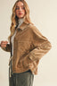 A woman wearing a cropped tan corduroy jacket over a white turtleneck and dark gray pants, photographed against a plain light beige studio background.