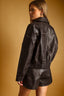 A woman wearing a dark brown leather jacket and matching leather shorts, shown from a side-back angle against a warm tan studio background.