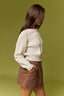 A woman shown from the back wearing a cream cable-knit sweater and brown shorts against a solid olive green background.