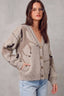A woman wearing a beige knit cardigan sweater with black abstract patterns and brown buttons, paired with light blue jeans, standing against a textured gray wall.