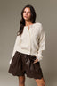 A woman modeling a cream-colored textured knit cardigan and dark brown faux-leather shorts against a solid taupe background.