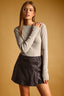 A woman modeling a light gray long-sleeve knit top and a dark gray faux-leather mini skirt against a warm tan studio background.