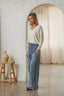A woman stands in a softly lit room wearing a cream V-neck long-sleeve sweater and light blue wide-leg jeans with dark side stripes. The setting features a textured white wall, a wooden stool with a ceramic jug, and a draped cream curtain in the background.