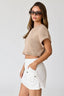 Woman wearing a beige cropped top and white shorts with sunglasses on a plain background