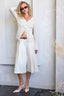 Woman in a white outfit standing against a light-colored wall.