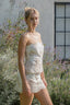 Woman in a white lace dress standing in a garden with flowers and a building in the background