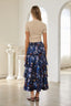 A woman shown from behind wearing a beige short-sleeve top and a long navy blue tiered maxi skirt with a colorful floral and botanical print, standing in a bright, white-walled room with arched windows.