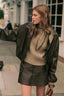 A woman wearing a dark olive brown faux-leather jacket, a beige knit sweater, and a matching dark brown faux-leather mini skirt, standing on a city street.
