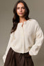 A woman wearing a cream waffle-knit button-front cardigan with a split neckline and dark brown satin-like skirt, photographed against a plain taupe studio background.