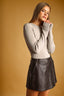 A woman modeling a light beige long-sleeve knit top and a dark brown faux-leather mini skirt against a warm tan studio background.