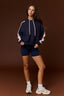 A woman modeling a navy blue hoodie with light pink sleeves and matching navy shorts, paired with white sneakers, against a solid dark brown studio background.