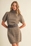 A woman modeling a taupe short-sleeve knit turtleneck dress with a wide dark belt, photographed against a plain light beige studio background.