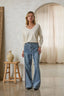 Woman wearing a white sweater and blue jeans standing in a room with a wooden stool and decorative vase.