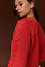 A close-up shot of a woman wearing a vibrant red cable-knit sweater, shown from a side-back angle against a dark brown studio background.