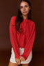 A woman wearing a bright red cable-knit sweater and white shorts, posed against a solid dark brown studio background.