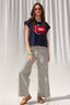 Woman wearing a navy blue sleeveless top with a red and white design and striped pants against a white curtain background.