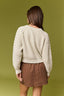 A woman shown from the back wearing a cream ribbed knit sweater and a brown mini skirt, standing against a solid olive green background.