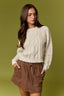 A woman wearing a cream cable-knit sweater and brown drawstring shorts against an olive green background.