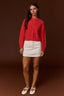 A woman stands against a dark brown studio background wearing a bright red cable-knit sweater, a white mini skirt, and white Mary Jane flats.