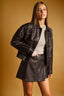 A woman modeling a black faux leather jacket and matching mini skirt set against a warm, solid mustard-yellow studio background.