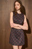 A woman modeling a sleeveless mini dress with a dark plaid pattern in black, burgundy, and brown tones, posed against a dark wood-paneled wall.