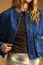 A close-up of a person wearing a cropped blue denim jacket over a dark brown top and a beige plaid skirt, against a warm tan background.