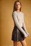 A woman modeling a light beige long-sleeve knit sweater paired with a dark brown faux-leather pleated mini skirt, set against a solid warm tan studio background.