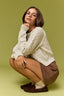 A woman crouching in a studio setting against a solid olive green background, wearing a cream cable-knit sweater, brown shorts, white socks, and dark brown loafers.