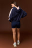 A woman shown from the back wearing a navy blue cropped hoodie with light pink sleeves, matching navy blue fitted shorts, white socks, and chunky white sneakers, set against a solid dark brown studio background.