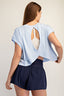 Woman wearing a light blue top with an open back and navy shorts on a beige background