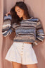 A woman wearing a fuzzy striped knit sweater with blue, brown, beige, and white tones, paired with white high-waisted shorts featuring gold buttons, posing against a dusty pink backdrop.