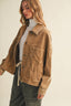 A woman wearing a light brown corduroy utility jacket over a white turtleneck and dark green pants, photographed against a plain off-white background.